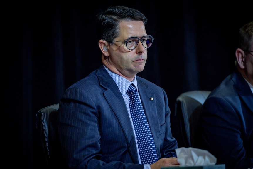 A man wearing glasses, short hair, and a blue suit sits on stage