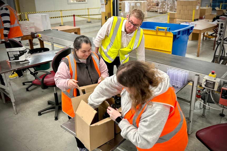 A middle-aged white man watches over two female workers packing items into boxes at a factory.