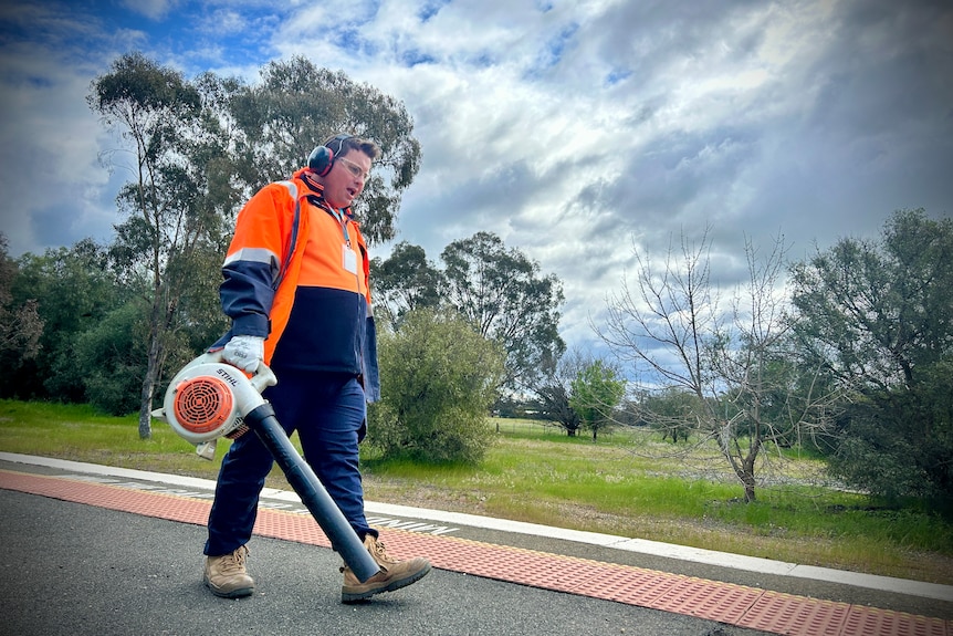 Caucasian man wearing orange hi-vis equipment using a leaf blower on a train platform